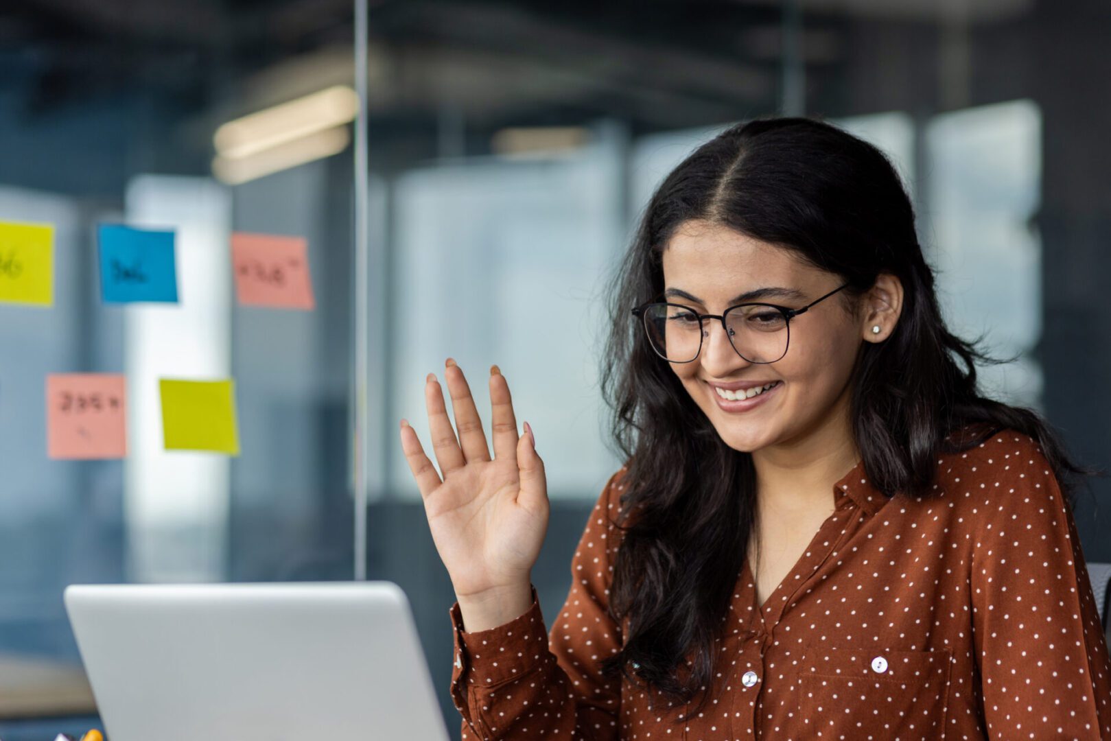 A woman with glasses waves while looking at her laptop.